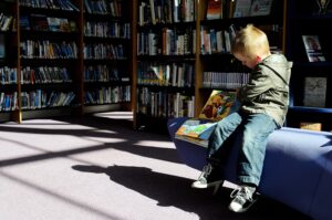 Young child sitting on bench in library scanning a book to his right side.+